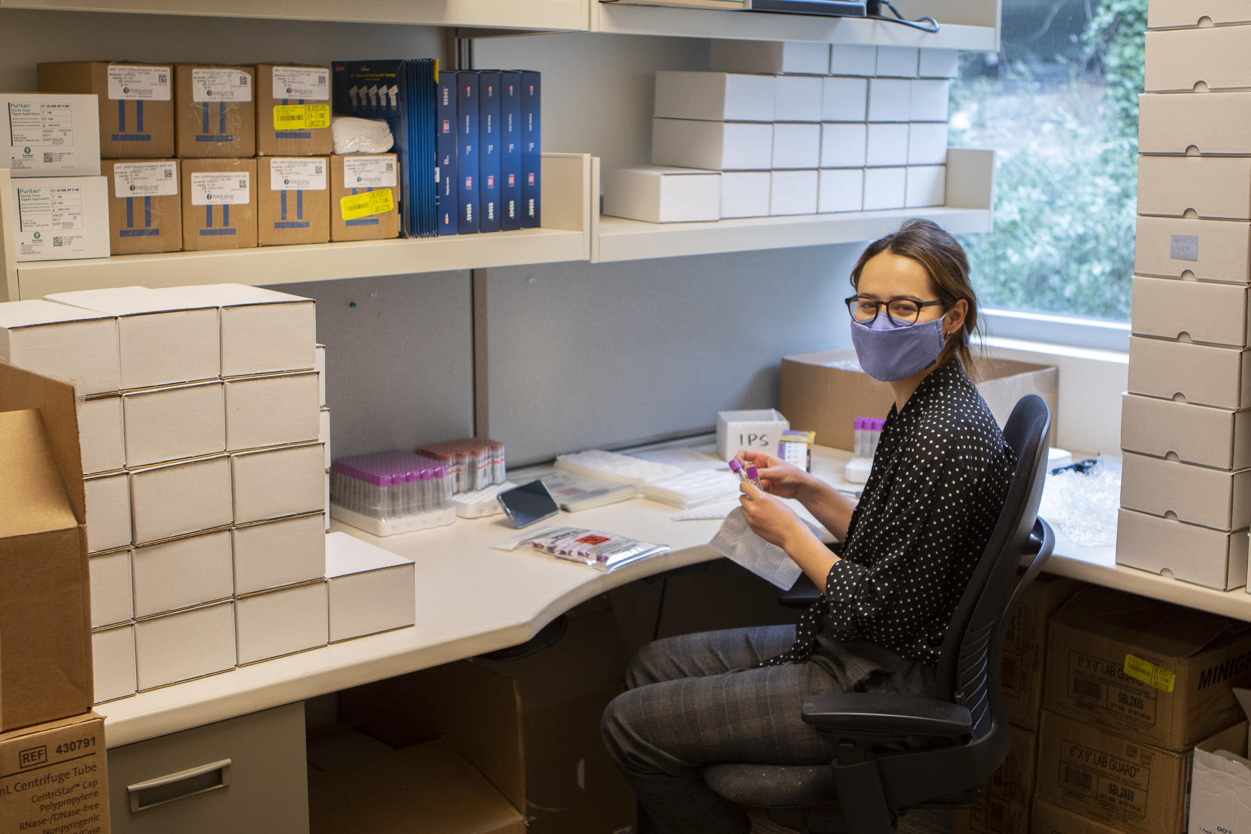 A woman is seated at a desk, surrounded by boxes and blood draw tubes.. She wears a mask and is turned toward the camera, eyes smiling.