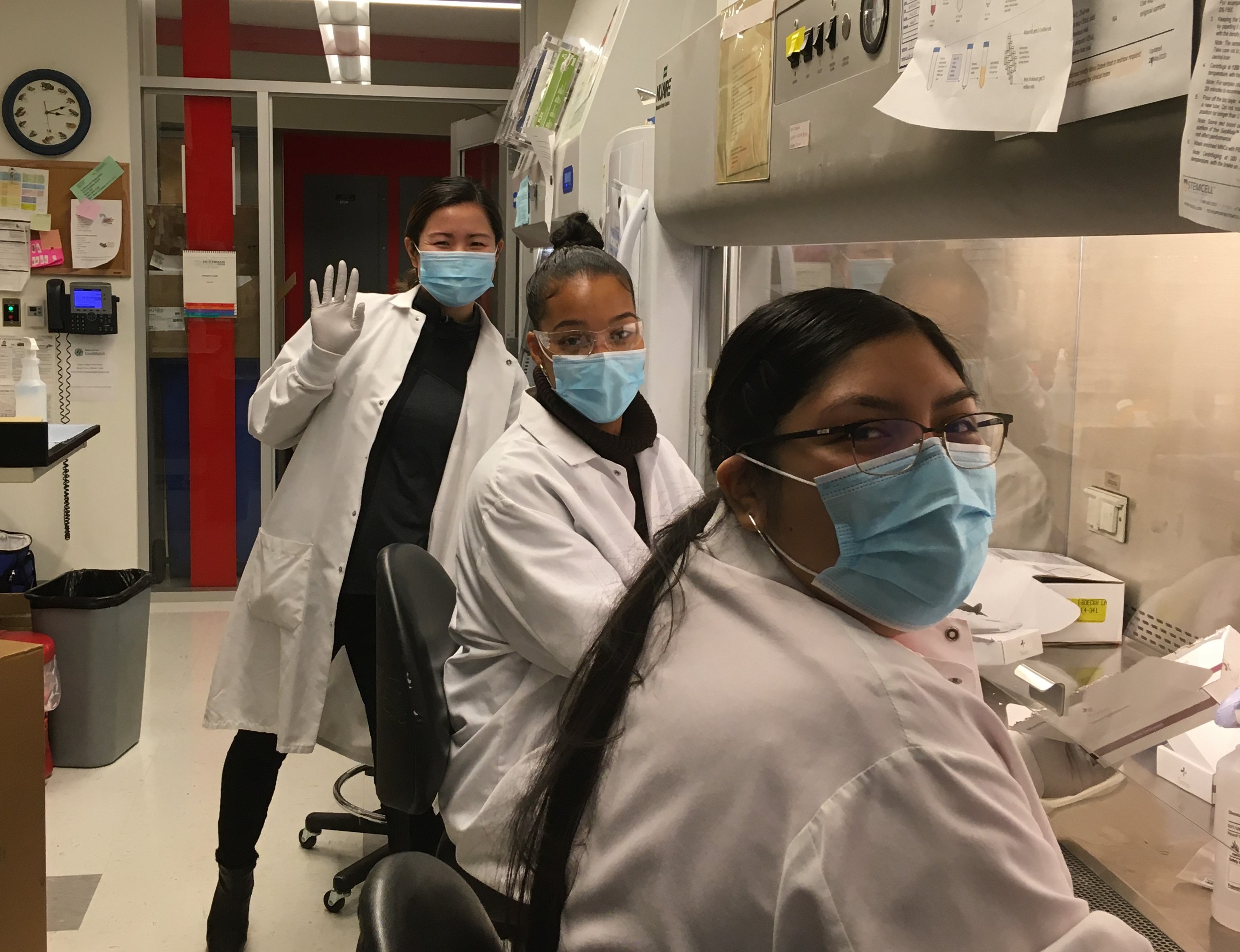 Three scientists in lab coats look at the camera and smile behind their masks