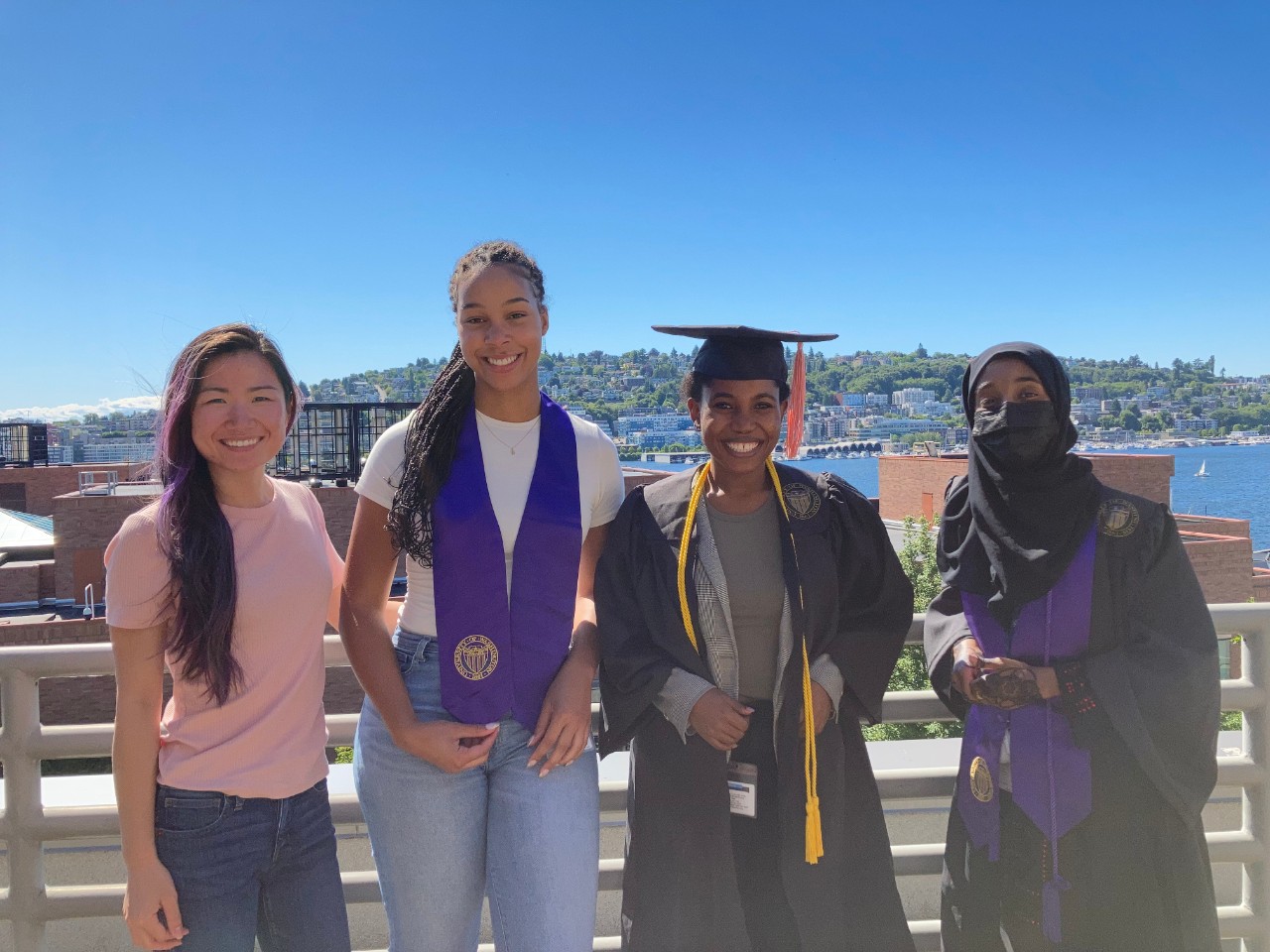 Four young women in graduation garments lean against a rail on a balcony on a sunny day